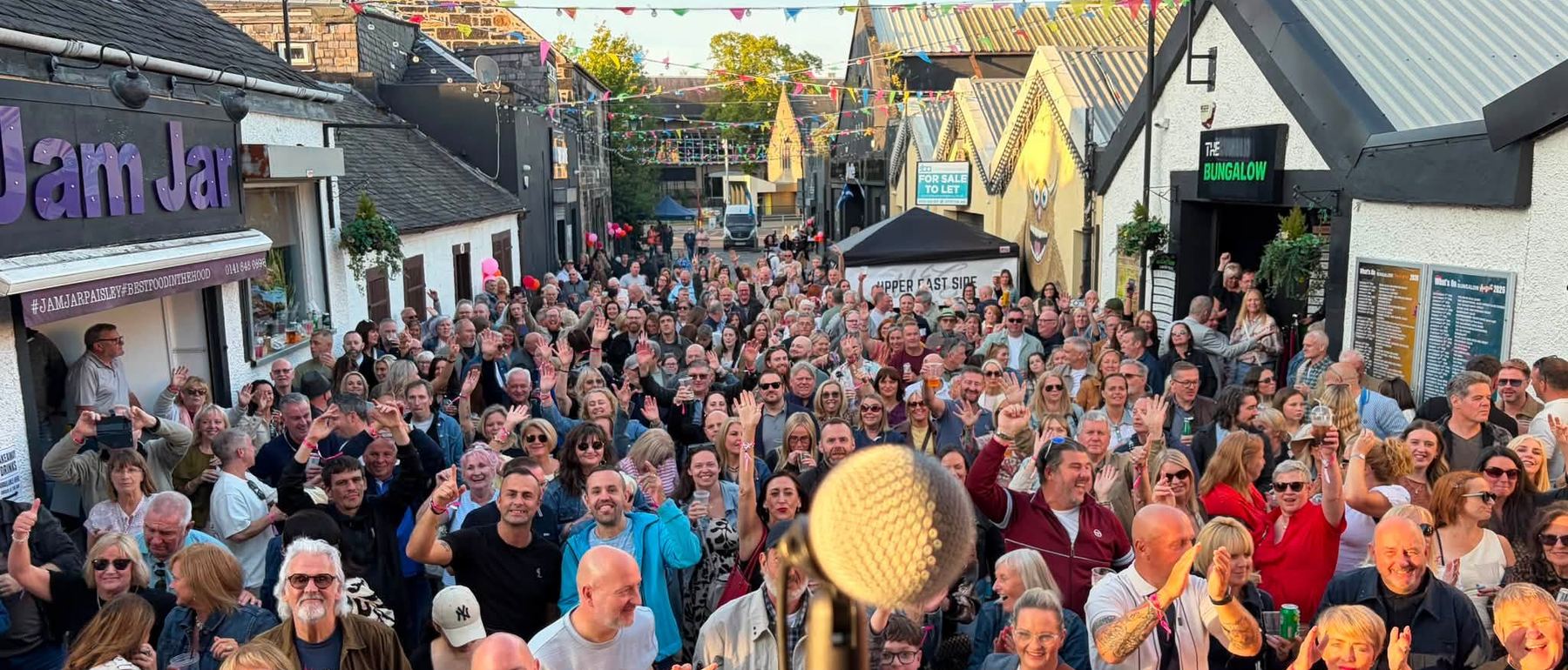 A packed Paisley street in the sunshine, hundreds of people with their hands in the air, seen past a microphone on the stage